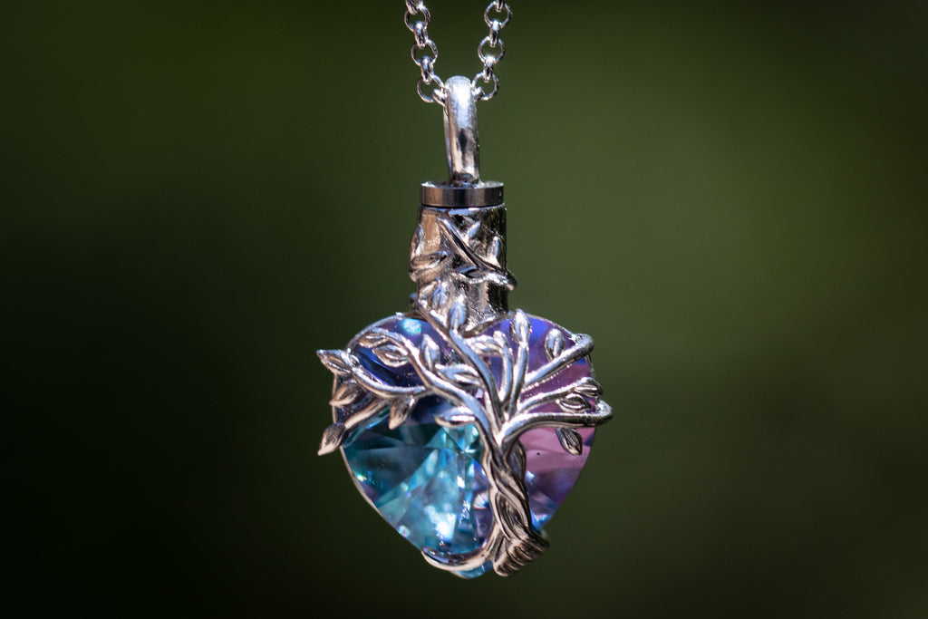 A heart-shaped pendant necklace featuring a faceted blue and purple iridescent gemstone wrapped in a silver tree design, hanging from a silver chain against a dark green blurred background.